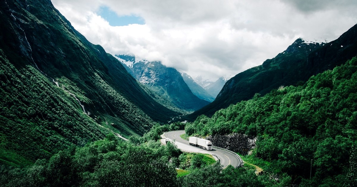 Vogntog kjører ned norsk fjellvei. Grønne lier. Fjell. 