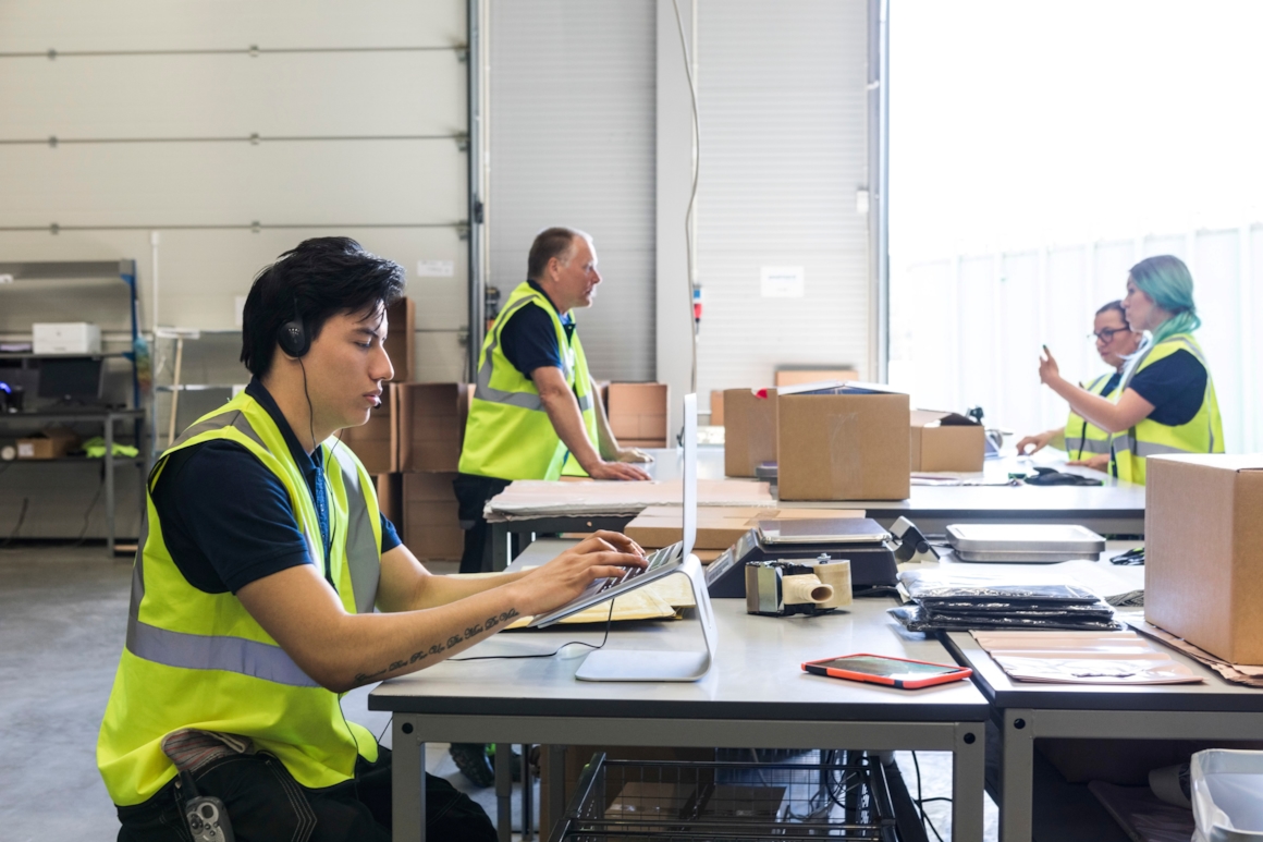fire kollegaer i gule refleksvester som jobber administrativt på et stort logistikklager